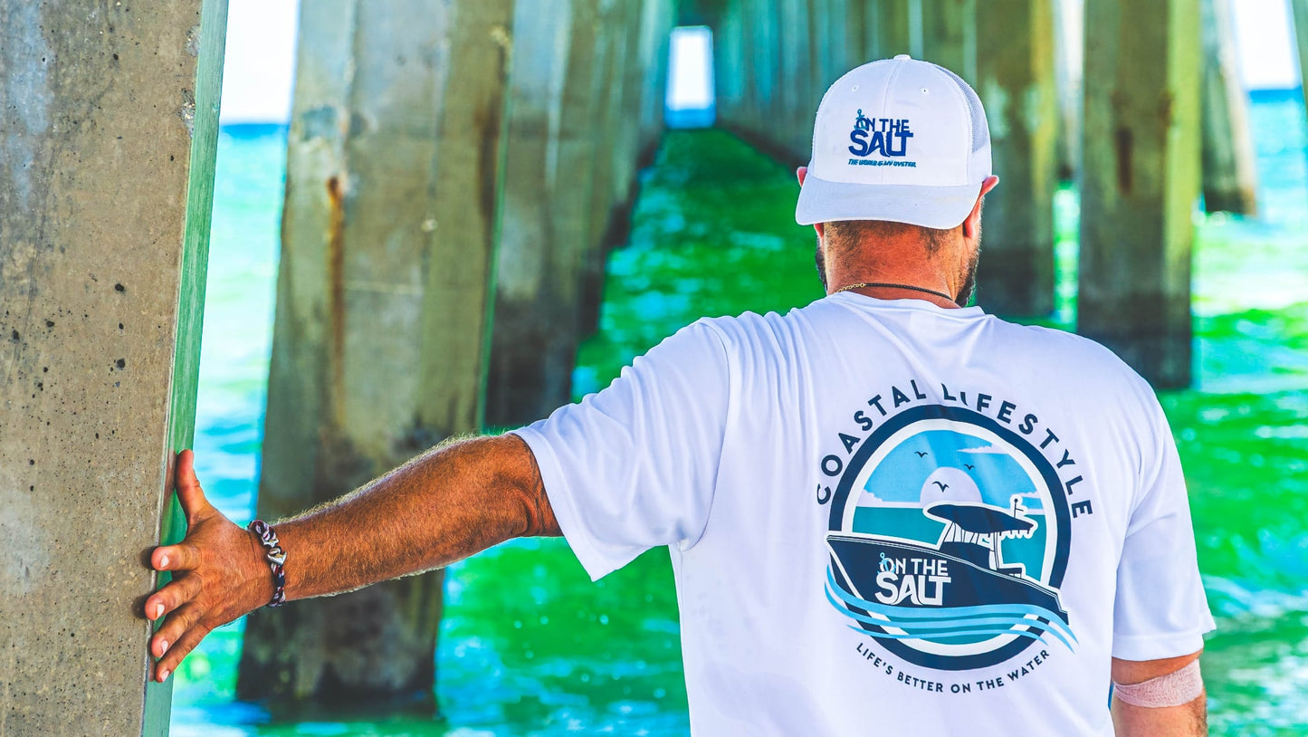 Man wearing a white t-shirt with 'Coastal Lifestyle' design, holding a surfboard by a pier.