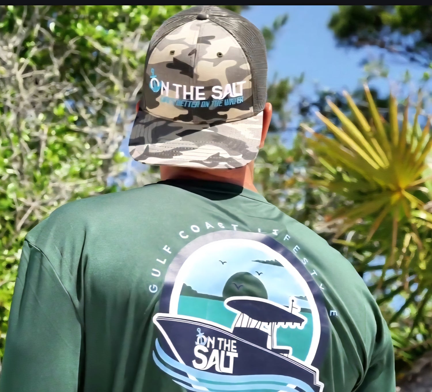A person wearing a green t-shirt with an embroidered design and a camouflage trucker hat with the text 'ON THE GO'.