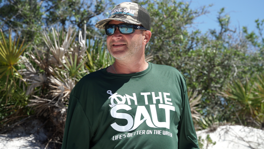 A person wearing a long sleeve green t-shirt with 'SALT' written across the front in white letters, standing on a beach.